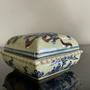 May include: A square ceramic trinket box with a lid. The box is decorated with a blue and white floral design and a brown and blue ribbon design. The box has a scalloped edge.