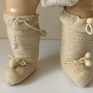 May include: A pair of white knitted doll boots with pom-poms on the toes and ties at the ankles.