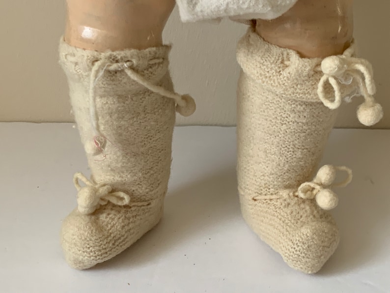 May include: A pair of white knitted doll boots with pom-poms on the toes and ties at the ankles.