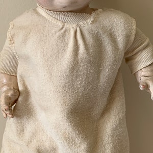 May include: A vintage porcelain doll wearing a white knitted gown with a ruffled neckline. The doll has a painted face and realistic features.