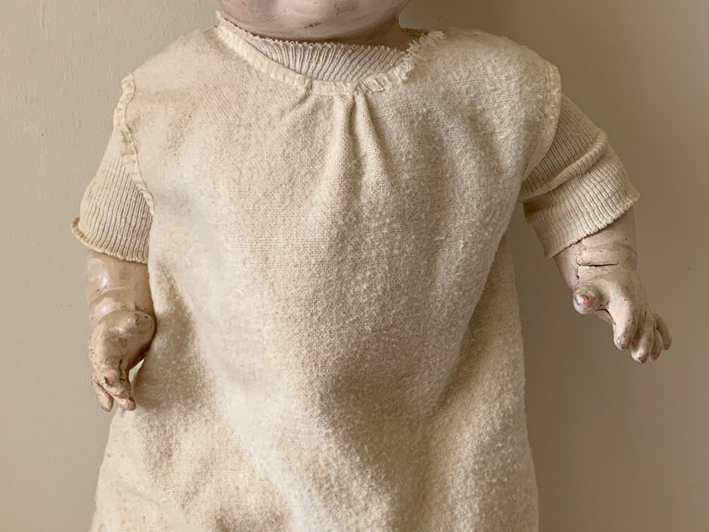 May include: A vintage porcelain doll wearing a white knitted gown with a ruffled neckline. The doll has a painted face and realistic features.