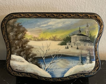 May include: A hand-painted wooden box with a winter scene. The box features a church with a white roof and a blue lake in the foreground. The box is decorated with gold trim and has the words "Палех" and "Тихонов" written on it.