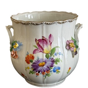 Puede incluir: Un jarrón de porcelana blanca con un borde festoneado y dos asas. El jarrón está decorado con flores pintadas a mano en tonos de rosa, amarillo, naranja, morado y azul. Las flores son tridimensionales y tienen una textura en relieve.