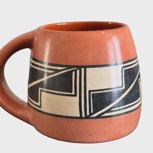 May include: A terracotta-coloured ceramic mug featuring a wide central band decorated with a geometric pattern of black and cream-coloured lines. The mug has a rounded body and a thick, curved handle extending from the side of the vessel.