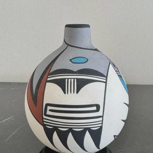 Op de afbeelding: Een bolvormige keramische vaas met een matte afwerking en een geometrisch ontwerp in zwart, wit, grijs, terracotta en turkoois. De vaas heeft een smalle hals en een rond lichaam versierd met gedurfde lijnen, hoekige vormen en gestileerde patronen.