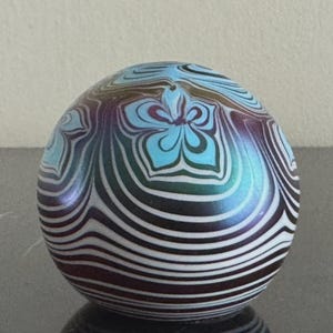 May include: A decorative glass orb with a swirling pattern of black and white stripes. The top features a light blue floral design. The orb has a reflective, iridescent finish, with hints of purple and green.