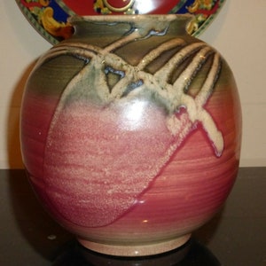 Puede incluir: Un jarrón redondo de cerámica con un esmalte marrón, verde y rosa. El jarrón tiene un diseño único con líneas y remolinos blancos.