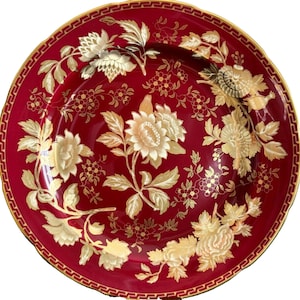 Op de afbeelding: Een ronde, dieprode plaat met een gouden bloemenontwerp. De plaat heeft een gouden rand en een zich herhalend geometrisch patroon langs de rand. Het bloemenontwerp toont grote bloemen en kleinere bloesems.