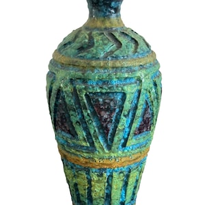 May include: A tall, textured ceramic vase with a narrow neck and a wide base. The vase features a geometric pattern in shades of teal, green, and brown, with accents of yellow. The surface has a rough, aged appearance, suggesting a vintage or handcrafted origin.
