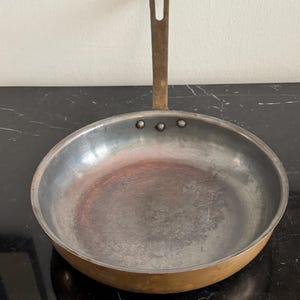 Op de afbeelding: Een gebruikte, ronde koekenpan met een lange, bronzen handgreep. De binnenkant van de pan is zilverkleurig met tekenen van slijtage en de buitenrand is warm brons. Klinknagels bevestigen de handgreep aan de pan.