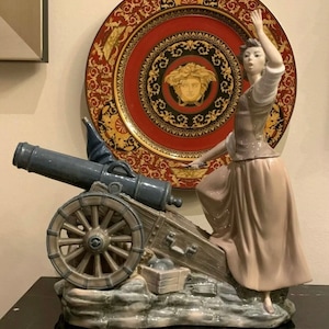 May include: A porcelain figurine of a woman in a white dress holding a cannon. The cannon is blue and grey and has a wooden wheel. The figurine is standing on a grey base with a brown wooden box and cannonballs.