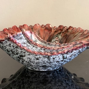 Op de afbeelding: Een decoratieve keramische kom met een gerimpelde rand. De kom heeft een gespikkelde grijze en witte buitenkant met een rood en bruin interieur. De rand heeft een golvend, getextureerd ontwerp en voegt visuele interesse toe aan het stuk.