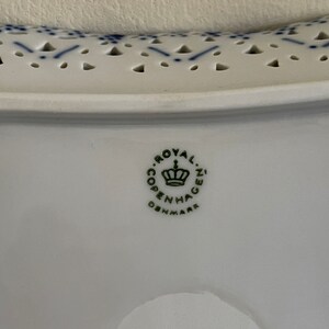 May include: Close-up of a white ceramic plate with a blue and white pattern. The plate has a scalloped edge and a hole pattern. The bottom of the plate has a green logo that reads "Royal Copenhagen Denmark".
