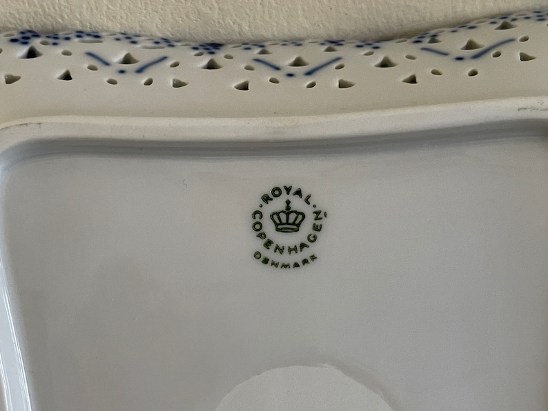 May include: Close-up of a white ceramic plate with a blue and white pattern. The plate has a scalloped edge and a hole pattern. The bottom of the plate has a green logo that reads "Royal Copenhagen Denmark".