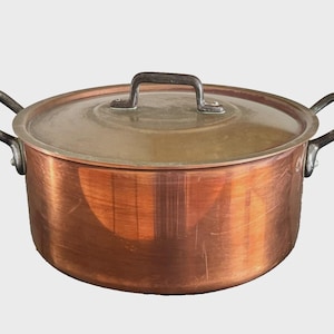 Op de afbeelding: Een ronde koperen pan met bijpassend deksel en twee donkere metalen handgrepen aan de zijkanten. De pan heeft een gepolijste koperen buitenkant met zichtbare horizontale korrel en een licht dof, mat metalen deksel met een handgreep.