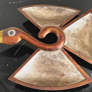 May include: A bird-shaped serving dish crafted from copper-toned metal with a polished brass beak and a dark circular eye inlay. The body features three distinct, triangular-shaped compartments with a textured, silver-toned interior finish.