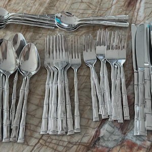 Könnte beinhalten: Eine Sammlung von Edelstahlbesteck, darunter Löffel, Gabeln und Messer. Das Besteck hat ein Bambus-Stil-Griffdesign. Die Utensilien sind auf einer gemusterten Oberfläche angeordnet und zeigen ein komplettes Set für das Essen.