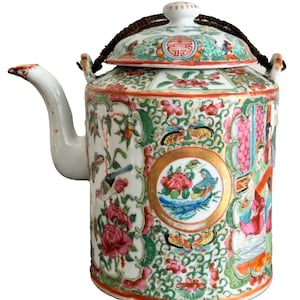 Puede incluir: Una tetera de porcelana blanca con un asa de cuerda marrón y una tapa decorada con un patrón floral en tonos de rosa, verde y dorado. La tetera tiene un cuerpo redondo y un pico en el lateral.