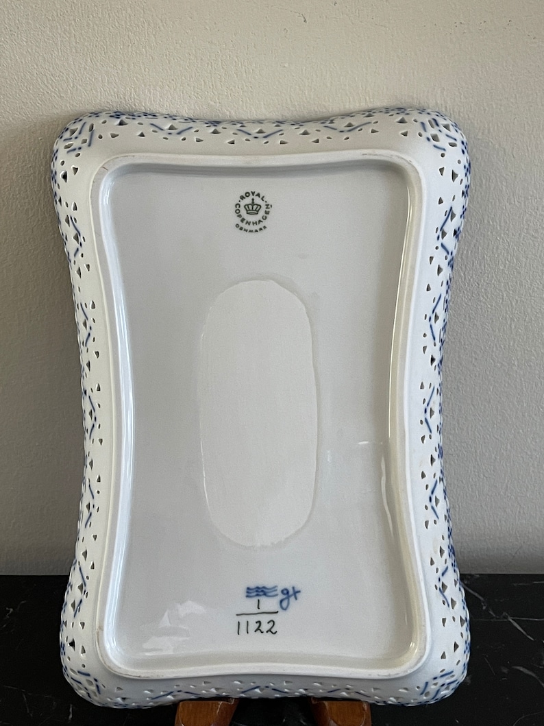 May include: A white ceramic serving dish with a blue and white pattern. The dish has a scalloped edge and a cut-out design. The bottom of the dish has the Royal Copenhagen logo and the number 1122.