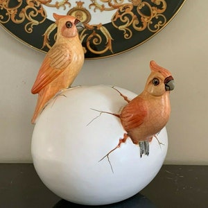 May include: Two orange and white ceramic cardinal birds, one perched on top of a large white egg with cracks, the other emerging from the egg.