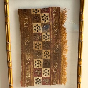 May include: A framed textile with a bamboo-style gold frame. The textile features a brown and tan color scheme with repeating square patterns containing stylized figures and floral motifs. The textile has a fringed edge.
