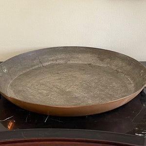 May include: A large, oval copper pan with two handles. The pan has a worn, aged patina and is likely vintage.