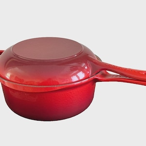 Puede incluir: Una cacerola de hierro fundido esmaltado rojo con tapa. La sartén tiene un mango largo y un pequeño asa de bucle en el lado opuesto. La batería de cocina es de un tono rojo vibrante.