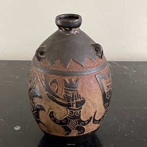 Può includere: Un piccolo vaso in ceramica marrone con corpo arrotondato e collo stretto. Il vaso è decorato con motivi geometrici neri e marroni e una figura stilizzata.