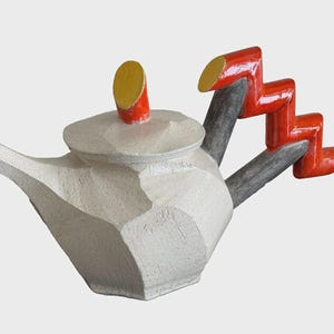 Könnte beinhalten: Eine weiße, geometrische Teekanne mit roten und gelben Akzenten an Griff und Deckel. Der Griff hat ein Zickzack-Design. Ausguss und Körper sind eckig, was ihr ein modernes, künstlerisches Aussehen verleiht. Die Teekanne ist ein einzigartiges Dekorationsstück.