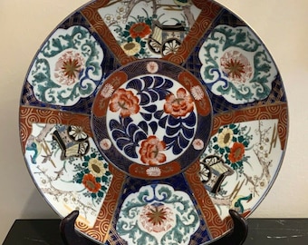 May include: A large, round, white ceramic plate with a blue, red, and gold floral design. The plate features a repeating pattern of stylized flowers, leaves, and geometric shapes. The plate is decorated with a gold rim and a gold band around the center.