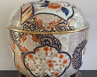 Puede incluir: Un gran jarrón de porcelana blanca con tapa, decorado con intrincados patrones florales y geométricos en oro, azul y rojo. El jarrón está sobre una base de madera.