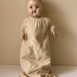 May include: Vintage porcelain baby doll with painted blue eyes, red lips, and rosy cheeks. The doll is wearing a white gown with a lace trim.