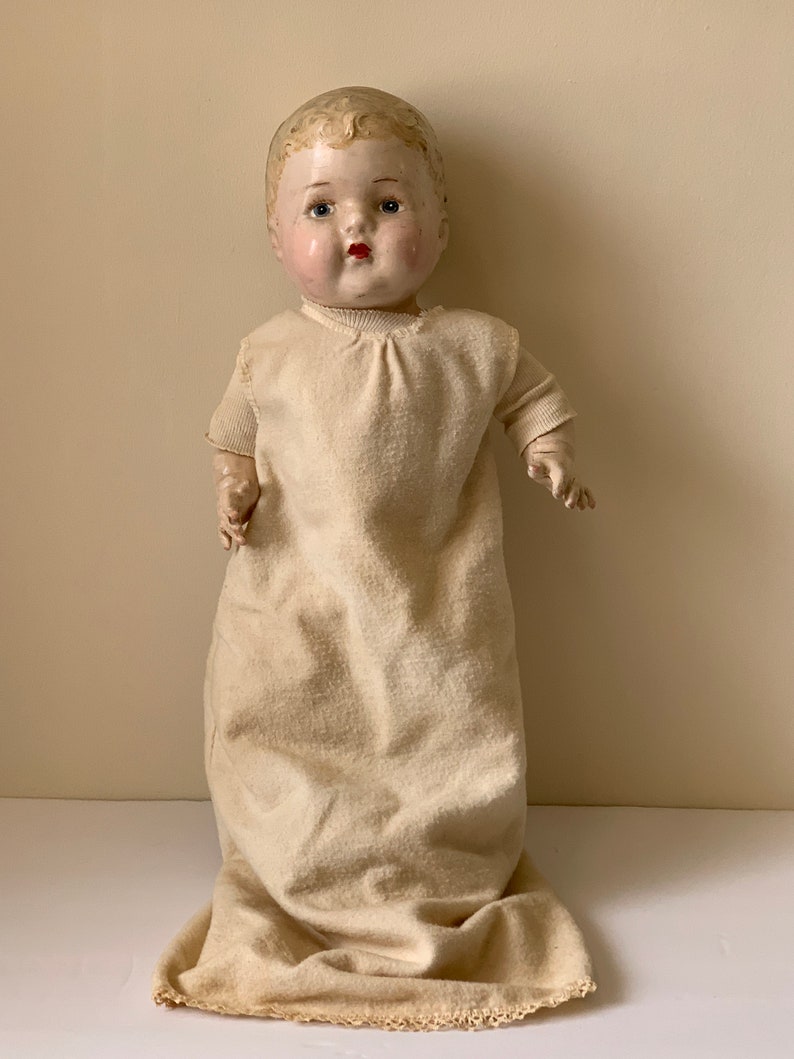 May include: Vintage porcelain baby doll with painted blue eyes, red lips, and rosy cheeks. The doll is wearing a white gown with a lace trim.
