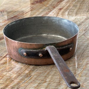 Puede incluir: Una cacerola de cobre vintage con un mango largo de hierro oscuro. La cacerola tiene un acabado de cobre martillado y tres remaches en el mango.