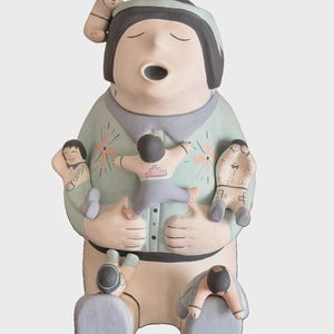 Pode incluir: Uma estatueta de cerâmica de um contador de histórias com uma figura central grande de olhos fechados e boca aberta, vestindo uma camisa verde-clara com padrões florais, rodeada por seis figuras menores presas ao corpo, pintadas em tons suaves de cinza, rosa e azul.