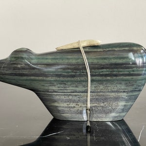 Op de afbeelding: Een groene en zwarte gestreepte stenen sculptuur van een beer met een wit bot op zijn rug. De sculptuur staat op een zwart oppervlak.