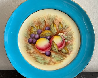 May include: A vintage ceramic plate with a blue rim and a gold trim. The center of the plate features a hand-painted design of fruit, including plums and peaches, on a cream background.