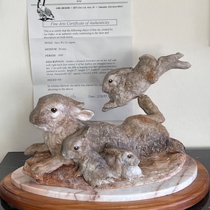 Puede incluir: Una escultura de bronce de una madre conejo con cuatro conejos bebés. La madre conejo está estirada de lado con una pata levantada. Los bebés están acurrucados junto a ella, con un bebé saltando sobre su pata extendida. La escultura está firmada "HALKO NWR" y titulada "Here We Go Again".