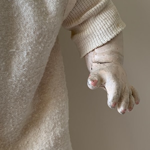 May include: Close-up of a vintage doll's hand, painted in a pale white with pink fingernails. The doll is wearing a white knitted sweater.