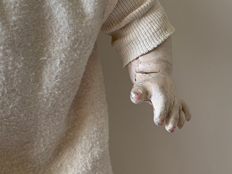 May include: Close-up of a vintage doll's hand, painted in a pale white with pink fingernails. The doll is wearing a white knitted sweater.