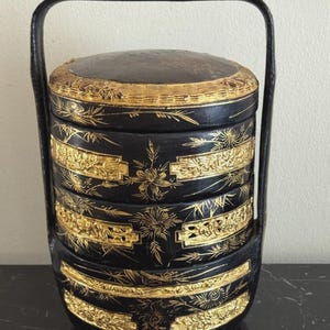 Op de afbeelding: Een zwarte, gelaagde, vintage voedseldrager met een handvat. De drager is versierd met gouden bloemen en geometrische ontwerpen. De bovenkant en banden rond elke laag zijn afgewerkt met goud. De handgreep is zwart.