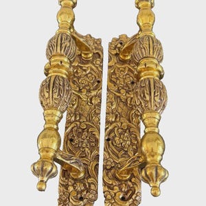 Peut inclure: Deux poignées de porte dorées et ornées. Chaque poignée comporte une longue plaque arrière rectangulaire avec des motifs floraux et des volutes complexes. Les poignées ont une forme bulbeuse avec des détails décoratifs et un levier. Le design est antique et élégant.