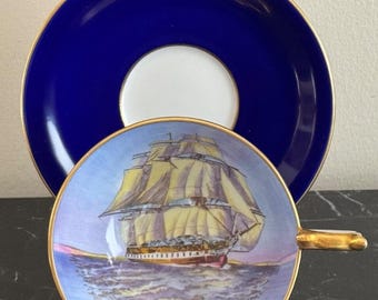 May include: A porcelain teacup and saucer set. The teacup features a detailed illustration of a sailing ship on the water, with yellow sails and a red hull. The saucer is a deep blue with a white center. Both pieces have gold trim.