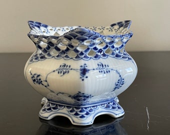 May include: A white porcelain sugar bowl with a blue and white floral pattern. The bowl has a scalloped edge and a lattice design on the top. The bowl is on a black surface.