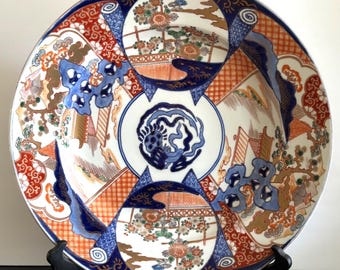 May include: A large, ornate ceramic plate with a detailed Japanese design. The plate features a central blue and white motif surrounded by orange, red, and blue geometric patterns and floral elements. The plate is displayed on a black stand.