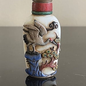 May include: A white porcelain snuff bottle with a green jade stopper. The bottle is decorated with a white crane in flight, perched on a branch with yellow flowers. The bottle has a blue and red base.