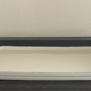 Könnte beinhalten: Rechteckige Keramik-Backform mit cremefarbenem Innenraum und dunkelblauem Rand. Die Form hat einen leicht erhöhten, gerippten Rand. Sie ist zum Backen und Servieren geeignet. Das Design ist schlicht und elegant.