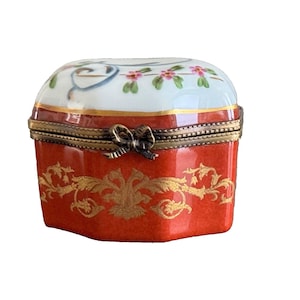 May include: A small, red porcelain trinket box with a gold floral design. The lid is white with a floral design in blue, green, and pink.