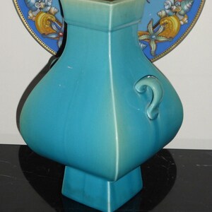 Pode incluir: Um vaso de cer&acirc;mica azul turquesa com base quadrada e corpo arredondado. O vaso tem uma al&ccedil;a de um lado e um acabamento liso e brilhante.