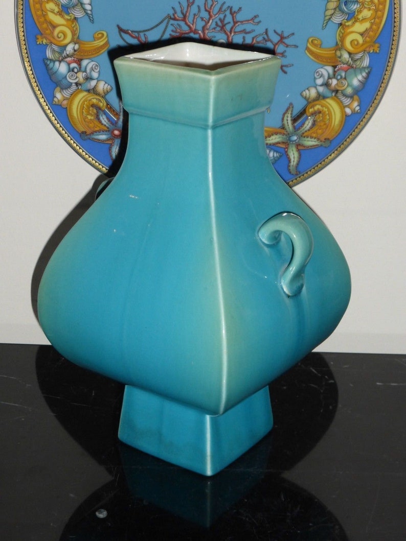Pode incluir: Um vaso de cer&acirc;mica azul turquesa com base quadrada e corpo arredondado. O vaso tem uma al&ccedil;a de um lado e um acabamento liso e brilhante.